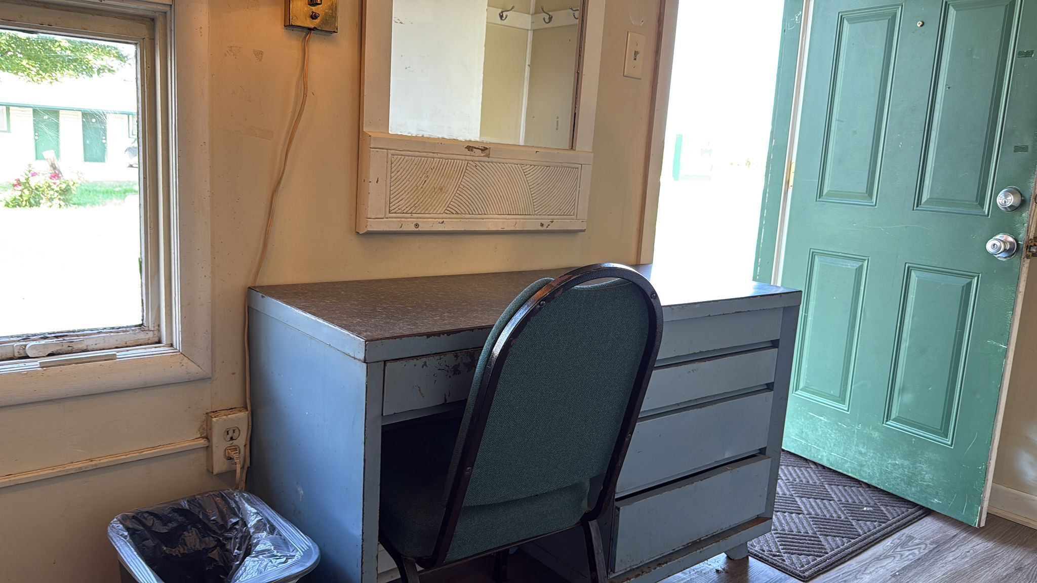 Een kamer met een bureau, een stoel, een raam, een deur en een afvalbak.