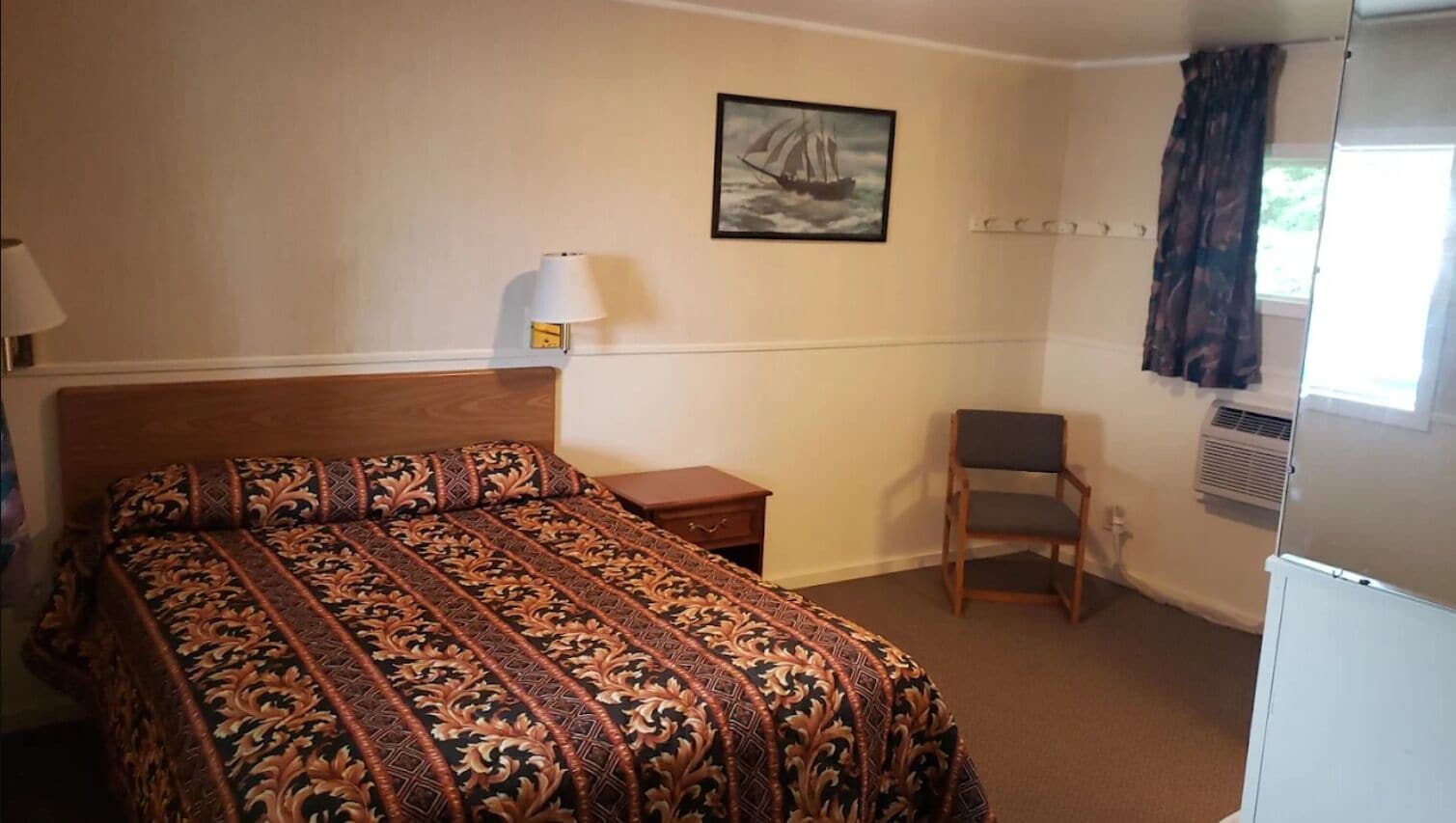 Une chambre d’hôtel avec un grand lit, une chaise, une petite table, une lampe, un tableau au mur et une fenêtre avec des rideaux.