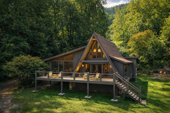 A modern two-story cabin with a deck, surrounded by dense forest.