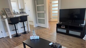 A modern living room with a dark wooden coffee table, a TV stand with a flat-screen TV, and a kitchen area with white cabinets and a bar stool.