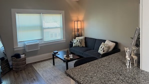A living room with a ceiling fan, a dark gray sofa, a coffee table, a window with blinds, a standing lamp, and a small decorative shelf.