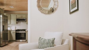 Un salón con un sillón blanco, un espejo redondo y una cocina al fondo.