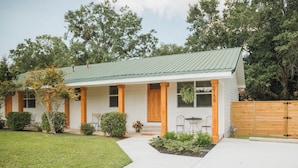 Una casa de dos pisos con techo de metal verde, paredes exteriores blancas y cerca de madera. Tiene un porche cubierto con mobiliario exterior y un pequeño jardín.