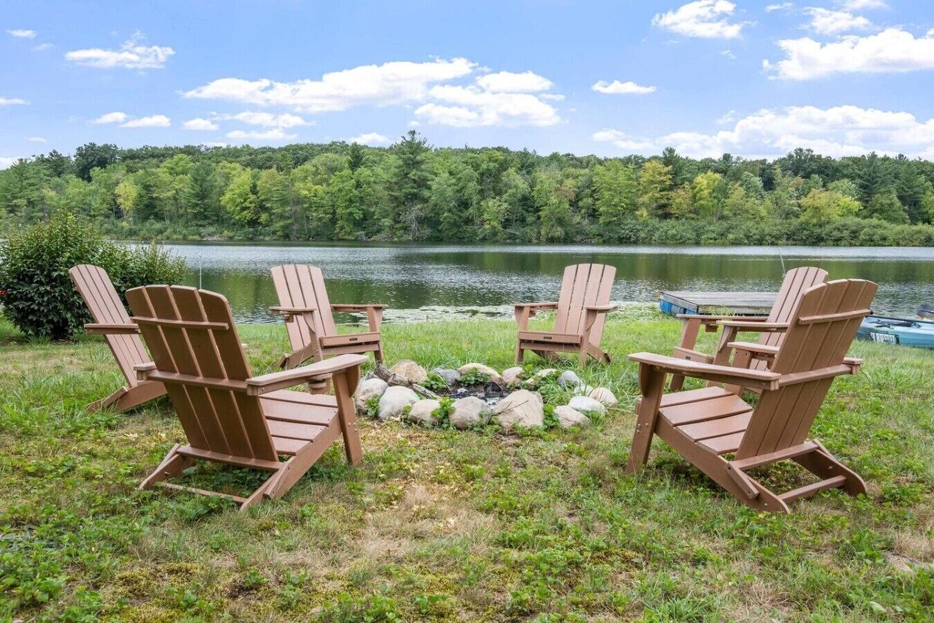 Four Adirondack chairs arranged around a campfire with a lake and forested hills in the background.