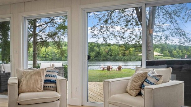 Una habitación con dos sillones color beige, una mesa de centro de madera y ventanas amplias con vistas a un lago y vegetación.