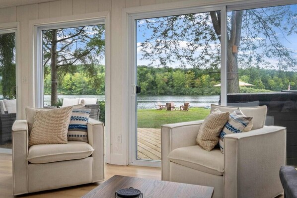A room with two beige armchairs, a wooden coffee table, and large windows offering a view of a lake and greenery.