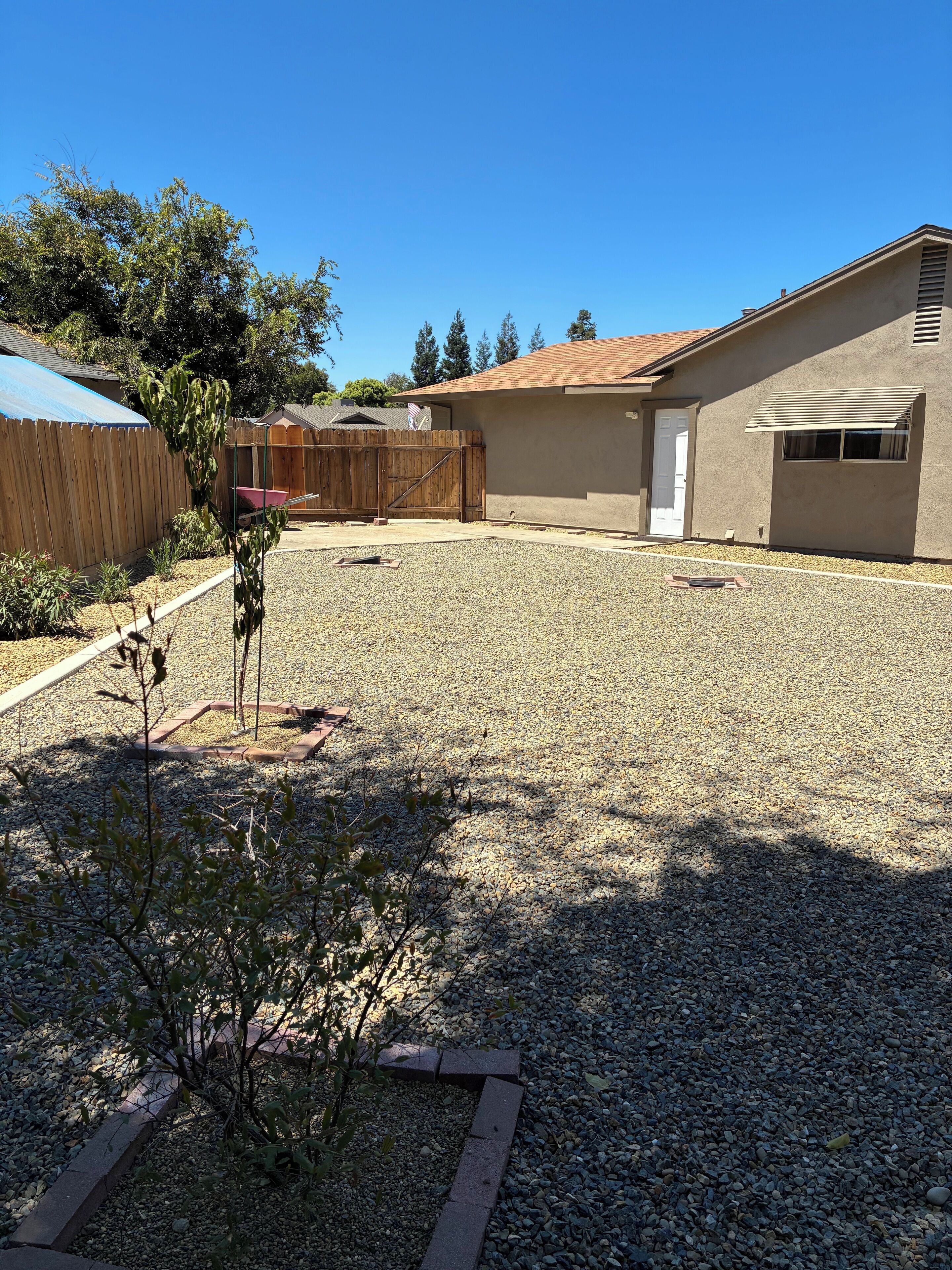 A residential backyard with a gravel driveway, a wooden fence, a small garden, and a house with a beige exterior and a red roof.