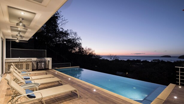 An infinity pool with lounge chairs, a barbecue area, and a view of the ocean at dusk.