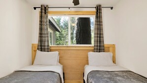 A room with two single beds, wooden headboards, a window with curtains, and a view of trees outside.