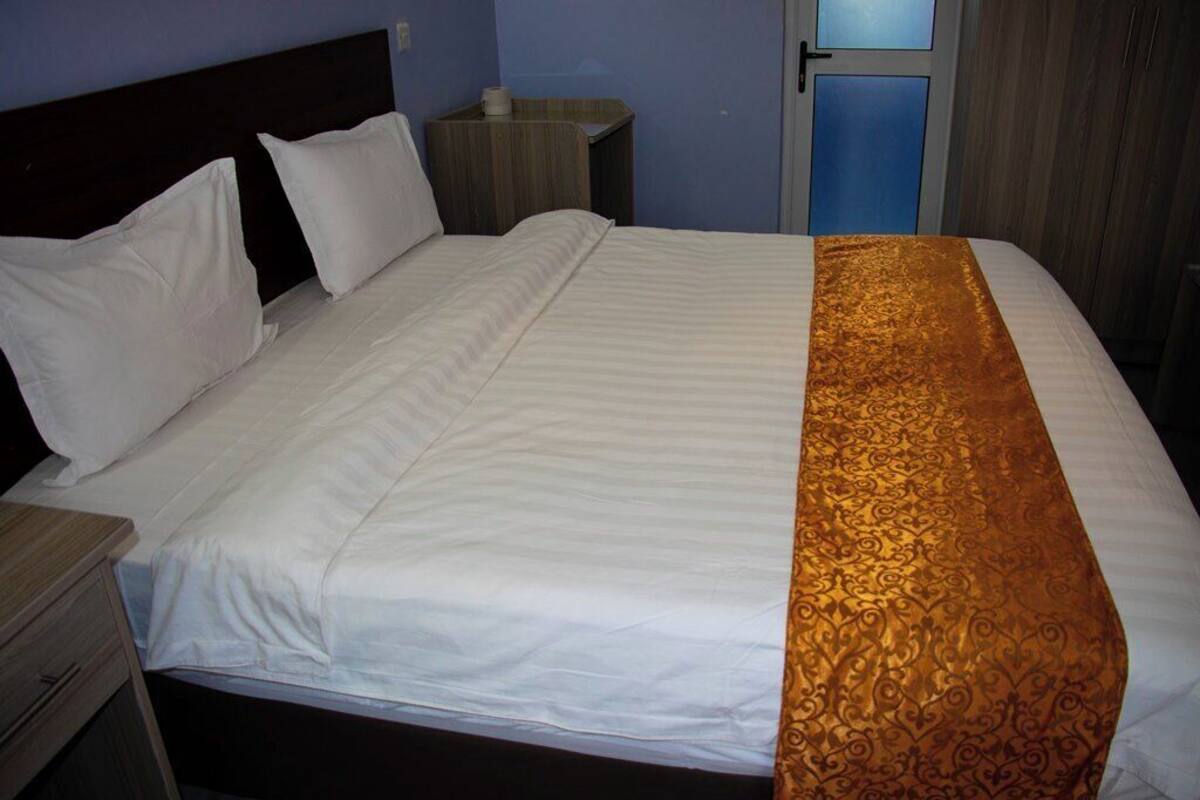 A neatly made bed with white linens and a decorative golden runner. There is a wooden nightstand with a lamp and a small table beside the bed.