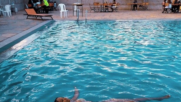 A swimming pool with a person floating. There are tables and chairs around the pool, and umbrellas providing shade. - Eastern Ark Hotel (Nguluni)