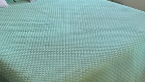 Un lit recouvert d’une couverture matelassée verte, avec une tête de lit blanche et une table de chevet sur laquelle se trouvent une lampe et une bouteille.