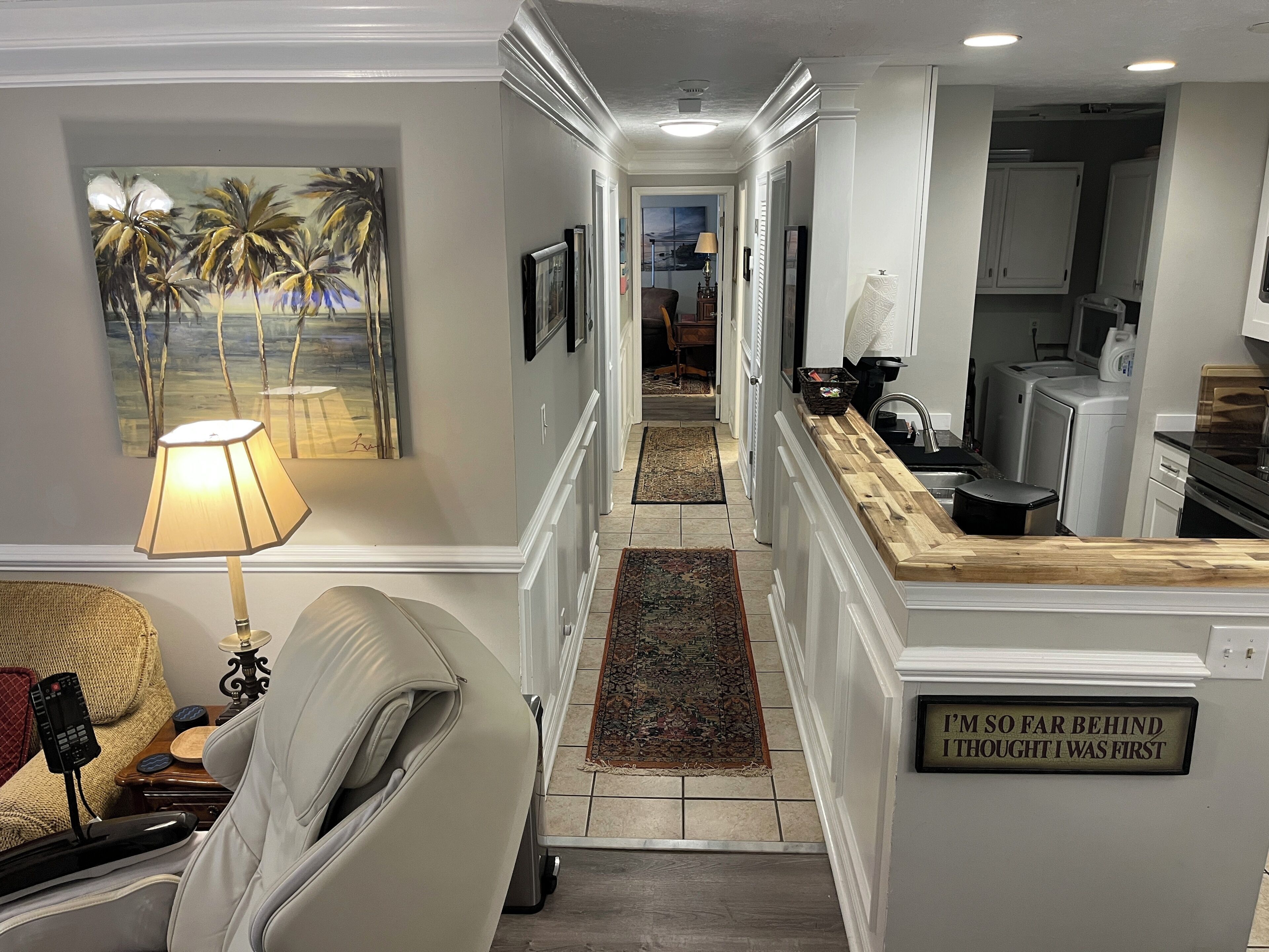 A hallway with a massage chair, a painting, and a kitchen area with a sign.