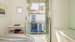 Un dormitorio con cama, una mesita de noche con libros, una ventana y vistas a una puerta azul y edificios.