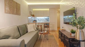 Ein modernes Wohnzimmer mit einer Couch, einem Couchtisch und einem Essbereich mit Stühlen. Im Hintergrund ist eine Küche mit Schränken und einer Arbeitsplatte zu sehen.