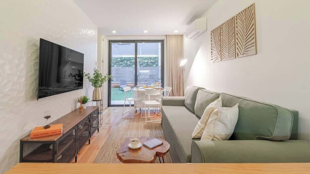 Ein modernes Wohnzimmer mit einem grünen Sofa, einem Holztisch, einem an der Wand montierten Fernseher und einem Balkon mit Gartenmöbeln.