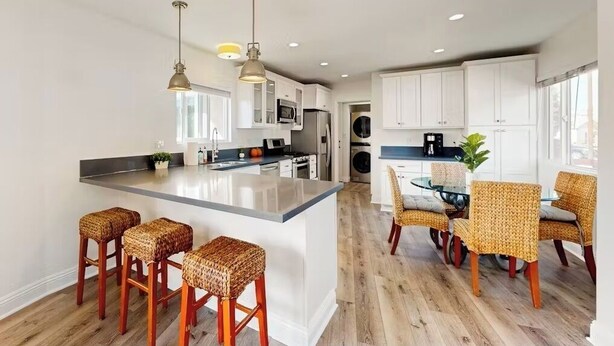 A modern kitchen with a central island, bar stools, a dining area with wicker chairs, and a view of the living room.