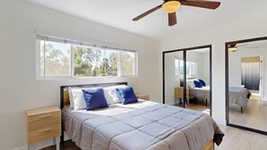 A bedroom with a bed, a nightstand, a ceiling fan, a large mirror, and a window with blinds.