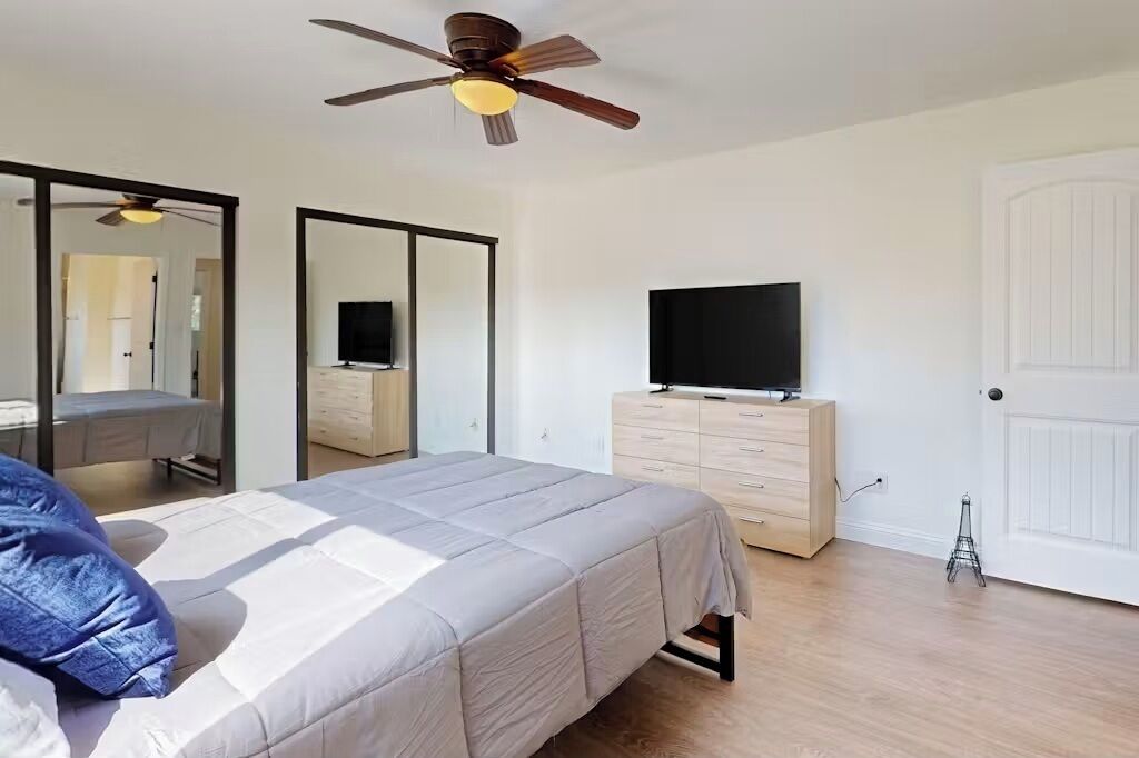 A bedroom with a bed, a TV on a dresser, a ceiling fan, and a door.