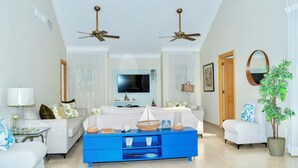 A modern living room with a blue sofa, white armchairs, a blue coffee table, and a television. There are ceiling fans, a large mirror, and a potted plant.