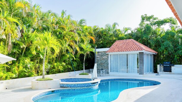 A swimming pool with a tiled edge, surrounded by lush greenery and a small building with a tiled roof.