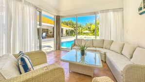 A spacious living area with a large glass door leading to a pool, wicker furniture, and a view of a tropical landscape.