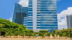 A tall, modern skyscraper with a glass facade, surrounded by greenery and under a clear blue sky.