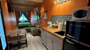 Eine kompakte Küche mit Mikrowelle, Spüle und Kühlschrank. Es gibt einen Essbereich mit Tisch und Stühlen. Der Raum hat eine Holzbalkendecke und orangefarbene Wände.