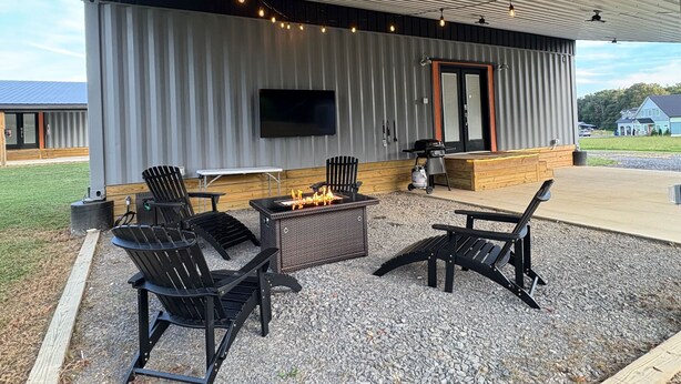Ein Außenbereich mit einer Terrasse, einem Feuerkorb, zwei Adirondack-Stühlen, einem Tisch und einem an der Wand befestigten Fernseher.
