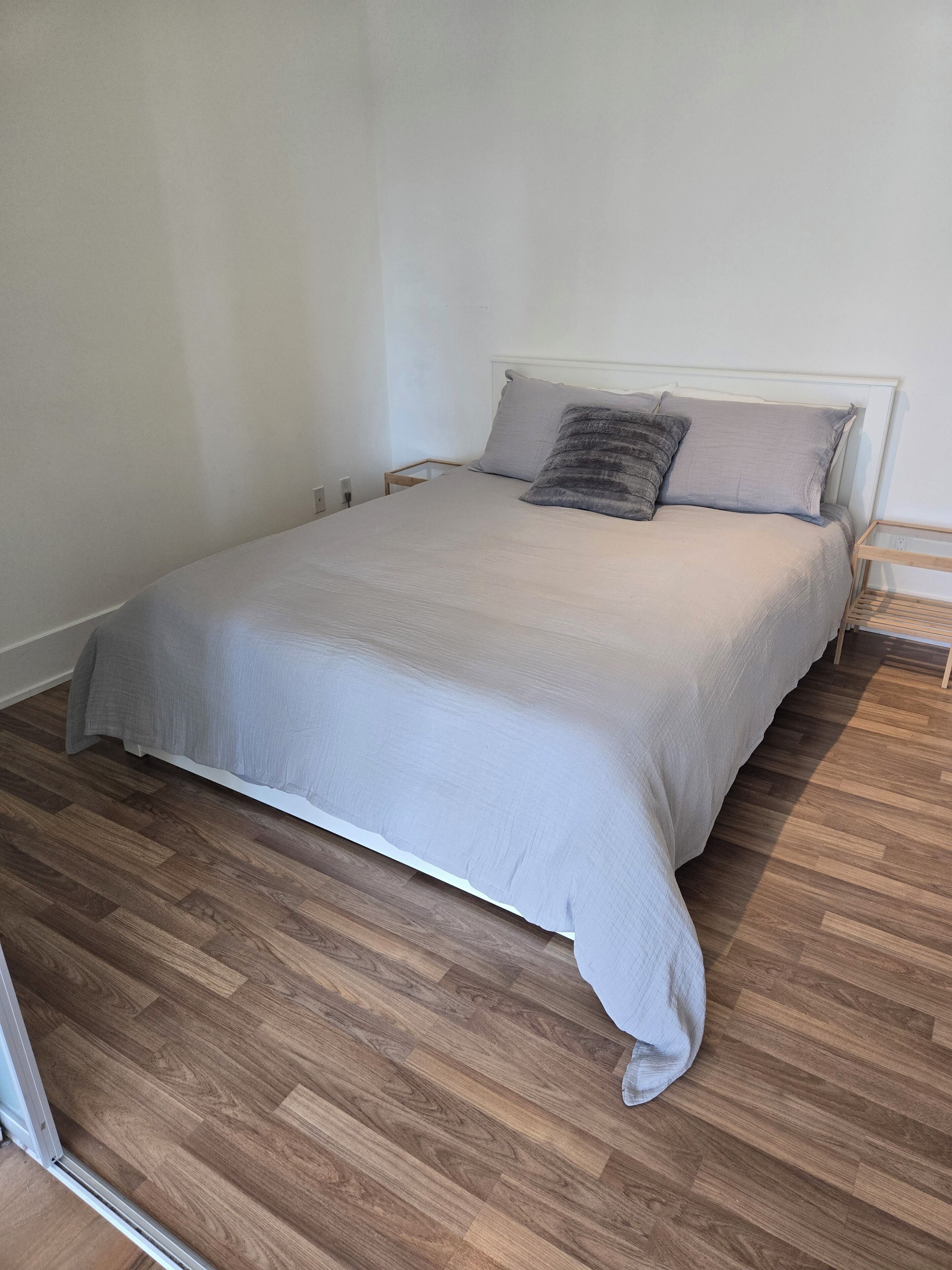A single bed with a grey bedspread and two pillows, a wooden nightstand, and a wooden floor.