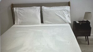 A bed with a metal frame and white bedding, a small bedside table with a lamp, and a plain wall in the background. - The palm trees (Las Vegas)