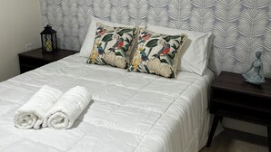 Een netjes opgemaakt bed met witte lakens, twee decoratieve kussens met een patroon van vogels en bloemen, een zwarte lantaarn op een nachtkastje, en een kleine sculptuur op een ander nachtkastje.