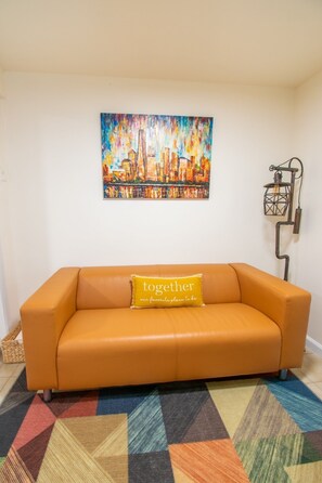 Quarto com sofá, tapete colorido, pintura de paisagem urbana e luminária fixada na parede.