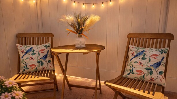 Twee houten stoelen met kussens met vogelmotief, een klein rond houten tafeltje en een vaas met gedroogde planten, alles voor een muur met lichtslingers.