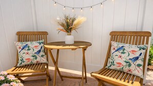 Twee houten stoelen met bloemkussens, een klein rond houten tafeltje en een vaas met gedroogde planten.