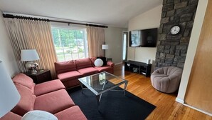 A modern living room with a red sofa, a glass coffee table, a grey armchair, a fireplace, and a TV mounted on a stone wall.