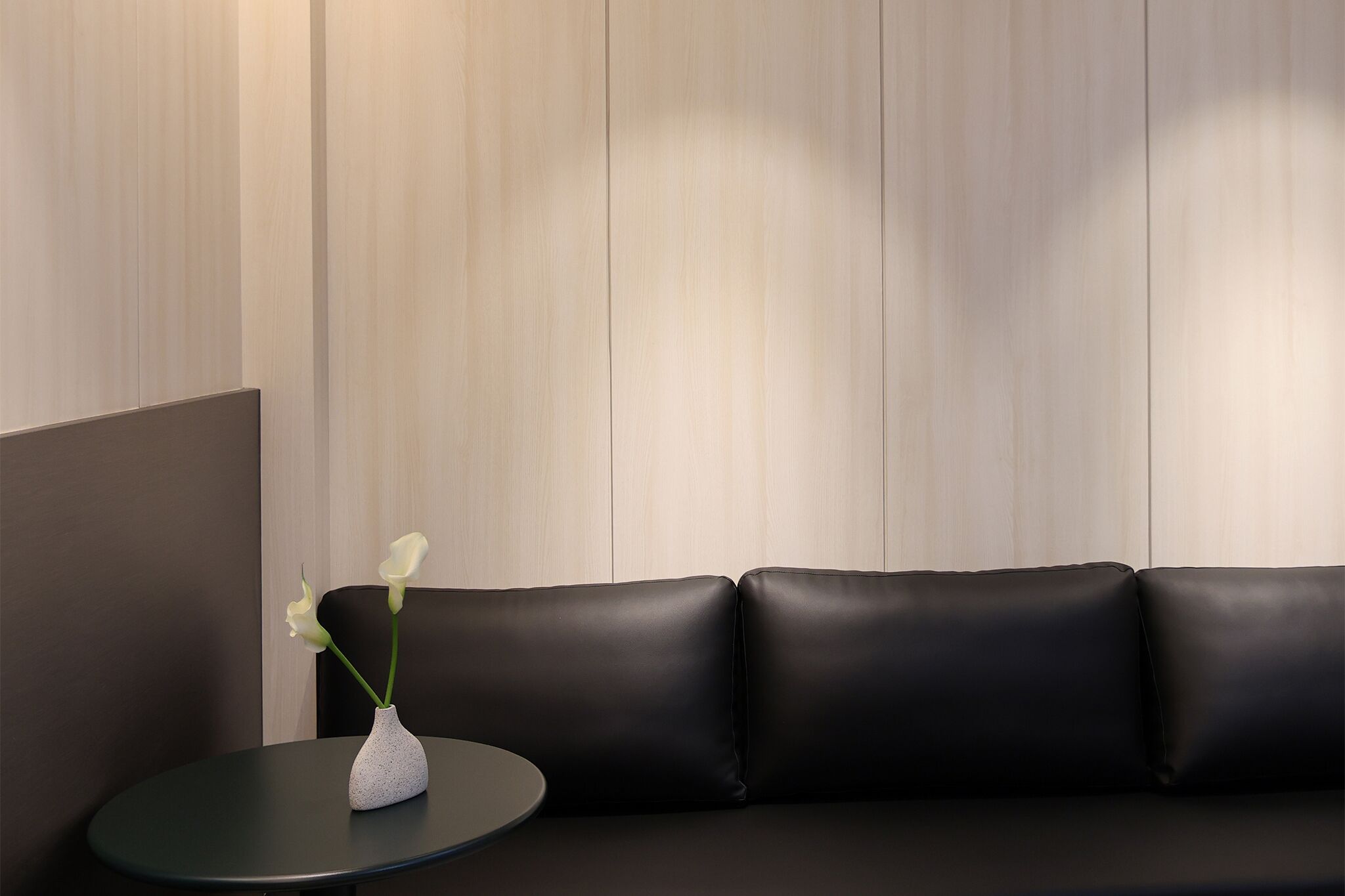 Un interior modern amb un sofà de pell negra, una taula rodona petita i un vas de flors blanc.