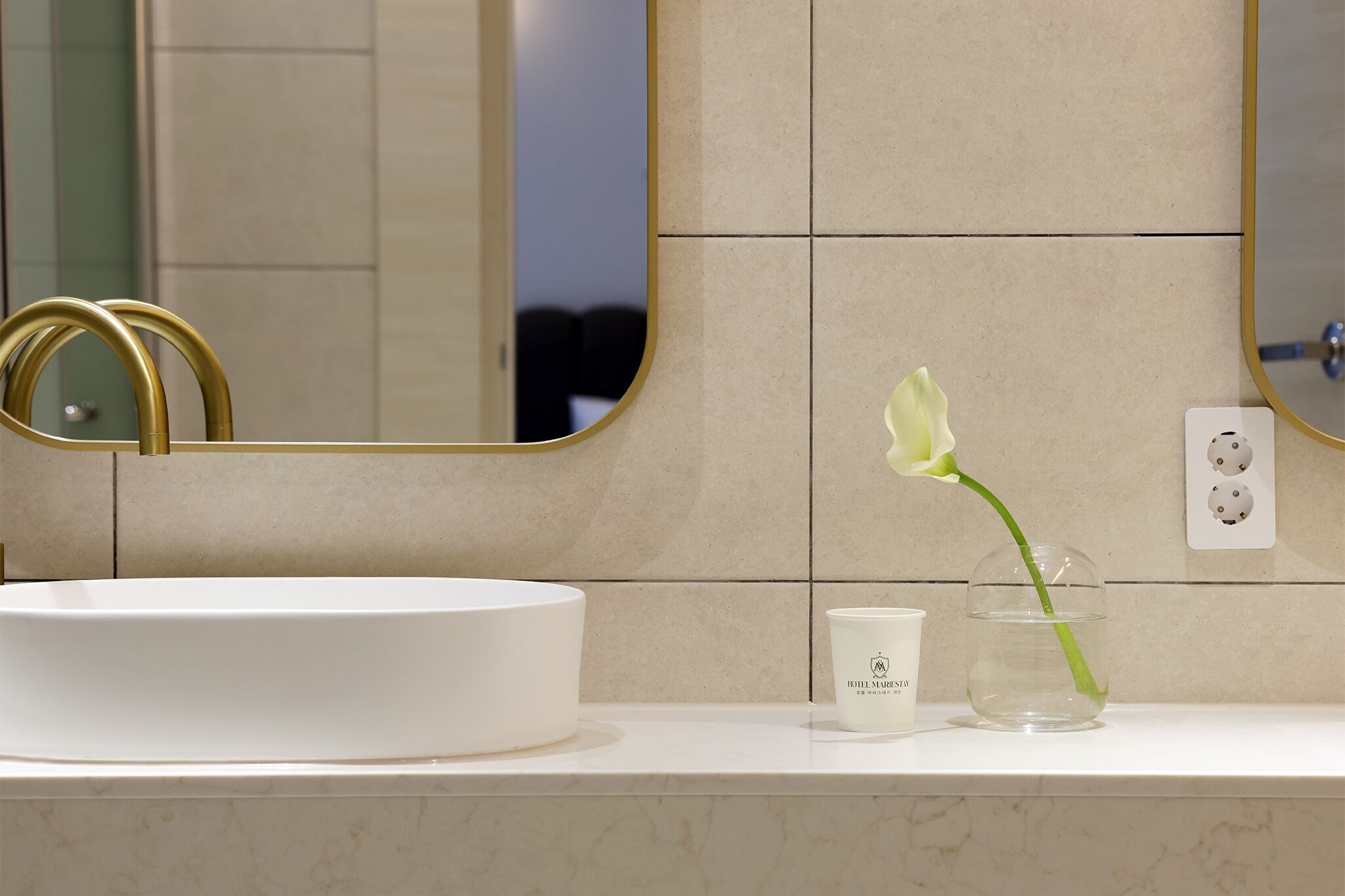 Un baño con un lavamanos blanco, un espejo con marco dorado y una flor blanca en un florero.