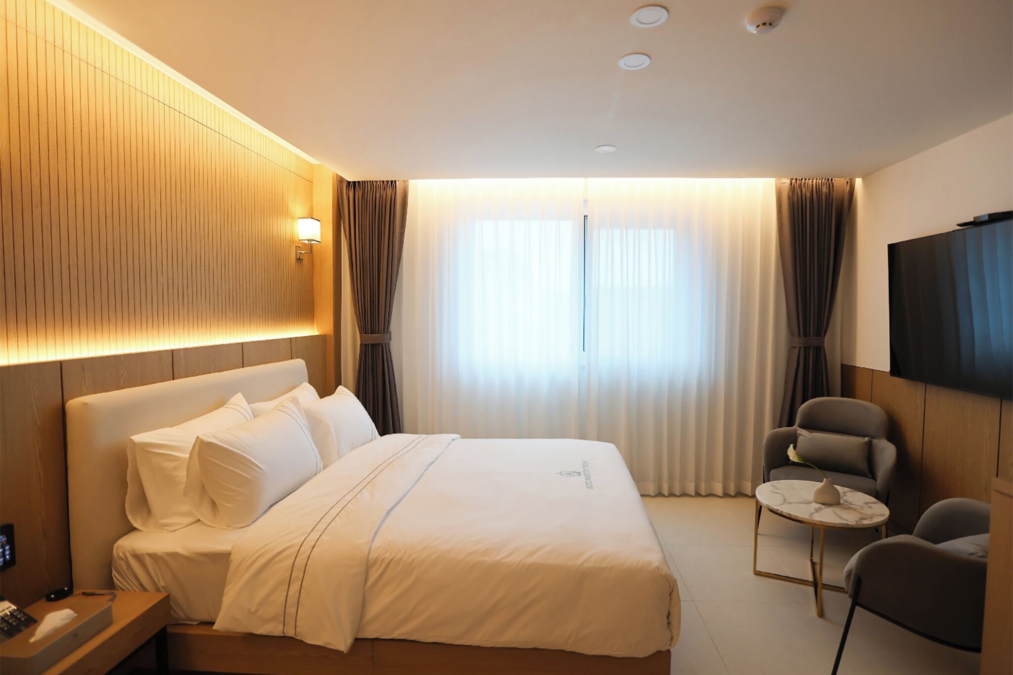 Quarto de hotel com uma cama, uma escrivaninha com telefone, uma cadeira, uma mesinha e uma televisão.