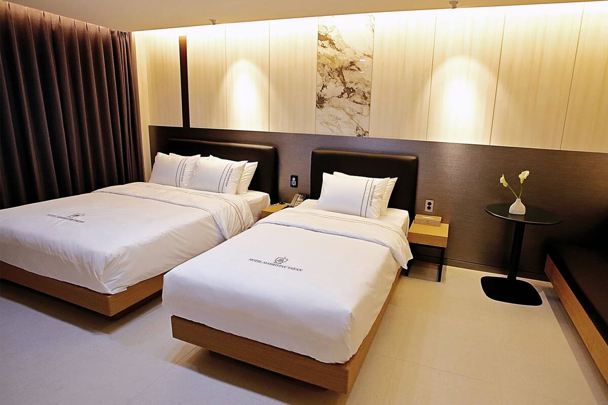 A hotel room with two beds, a nightstand, a small table with a vase, and a large window with curtains.