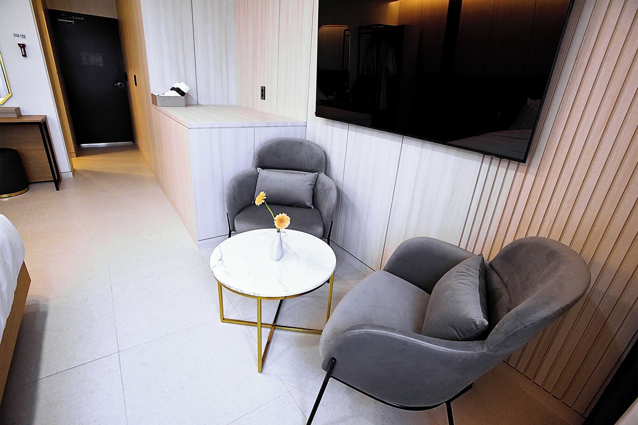 Une chambre d’hôtel moderne avec un lit, une petite table ronde ornée d’un vase de fleurs, deux fauteuils gris et un grand téléviseur à écran plat fixé au mur.