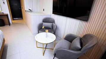 Una habitación de hotel moderna con una cama, una pequeña mesa redonda con un jarrón de flores, dos sillones grises y un televisor de pantalla plana grande montado en la pared.