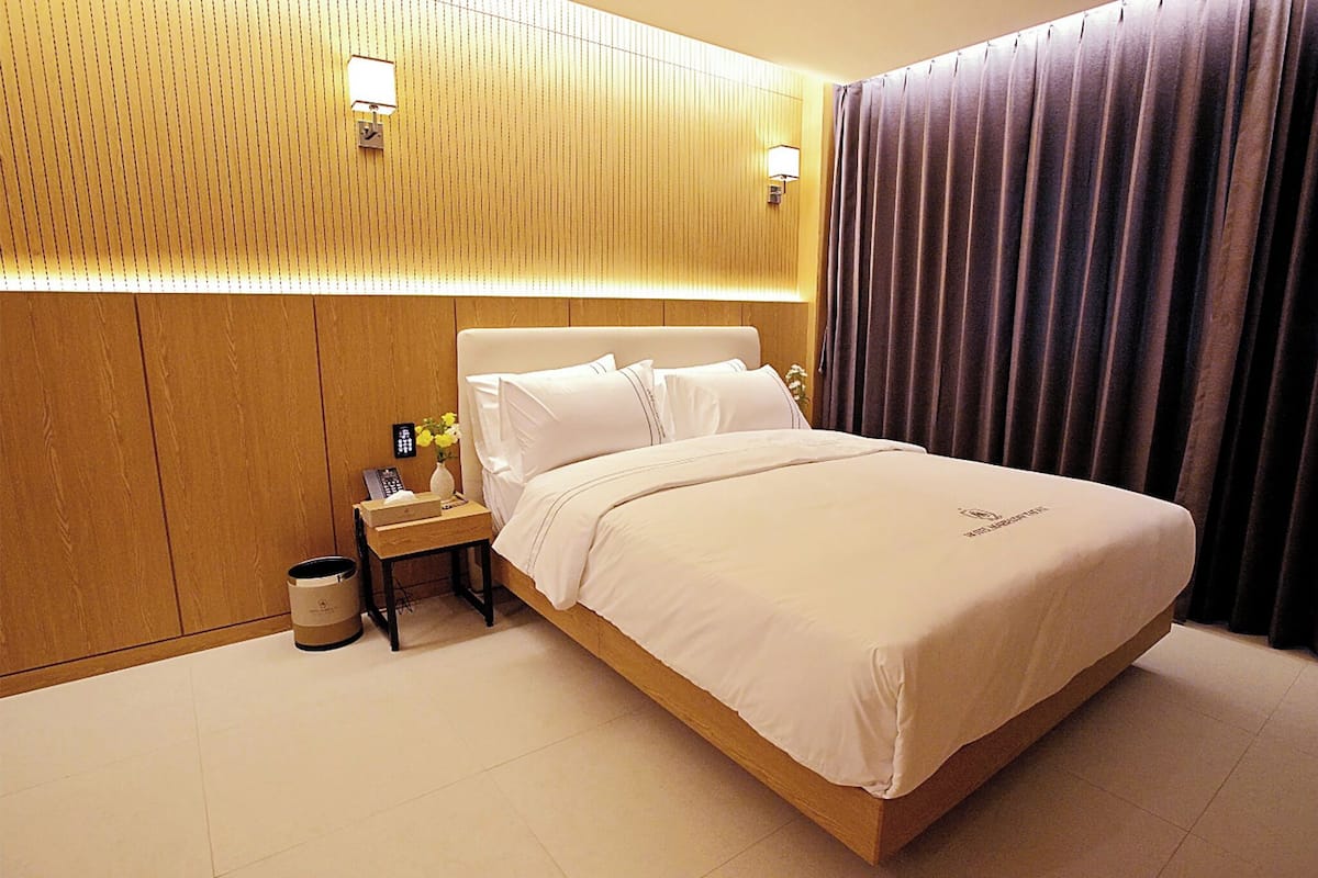 A hotel room with a wooden headboard, a bedside table with a lamp, and a large bed with white bedding.
