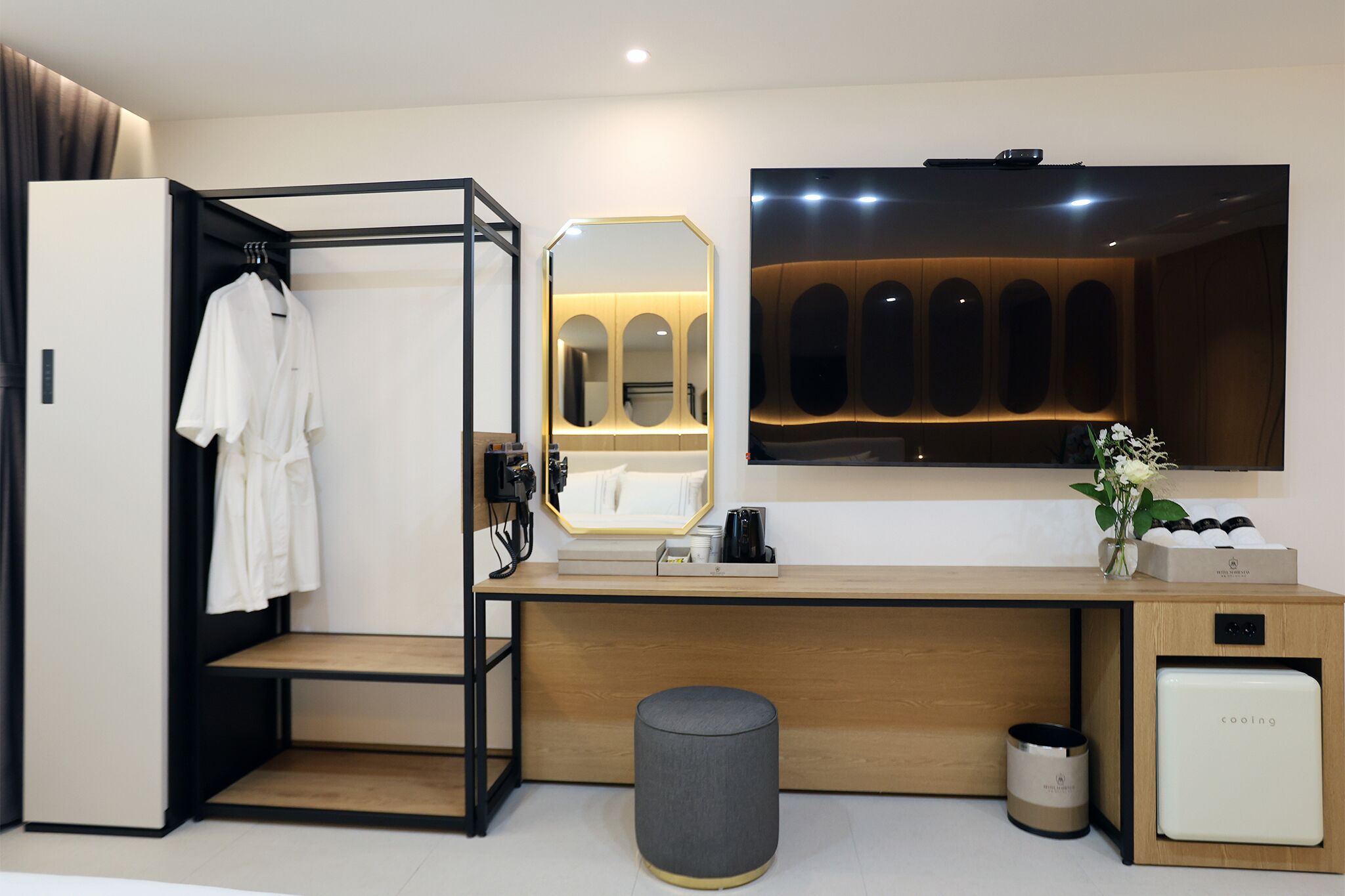Ein modernes Hotelzimmer mit Schreibtisch aus Holz, integriertem Spiegel, kleinem Kühlschrank, Schminktisch mit Spiegel und Kleiderständer.