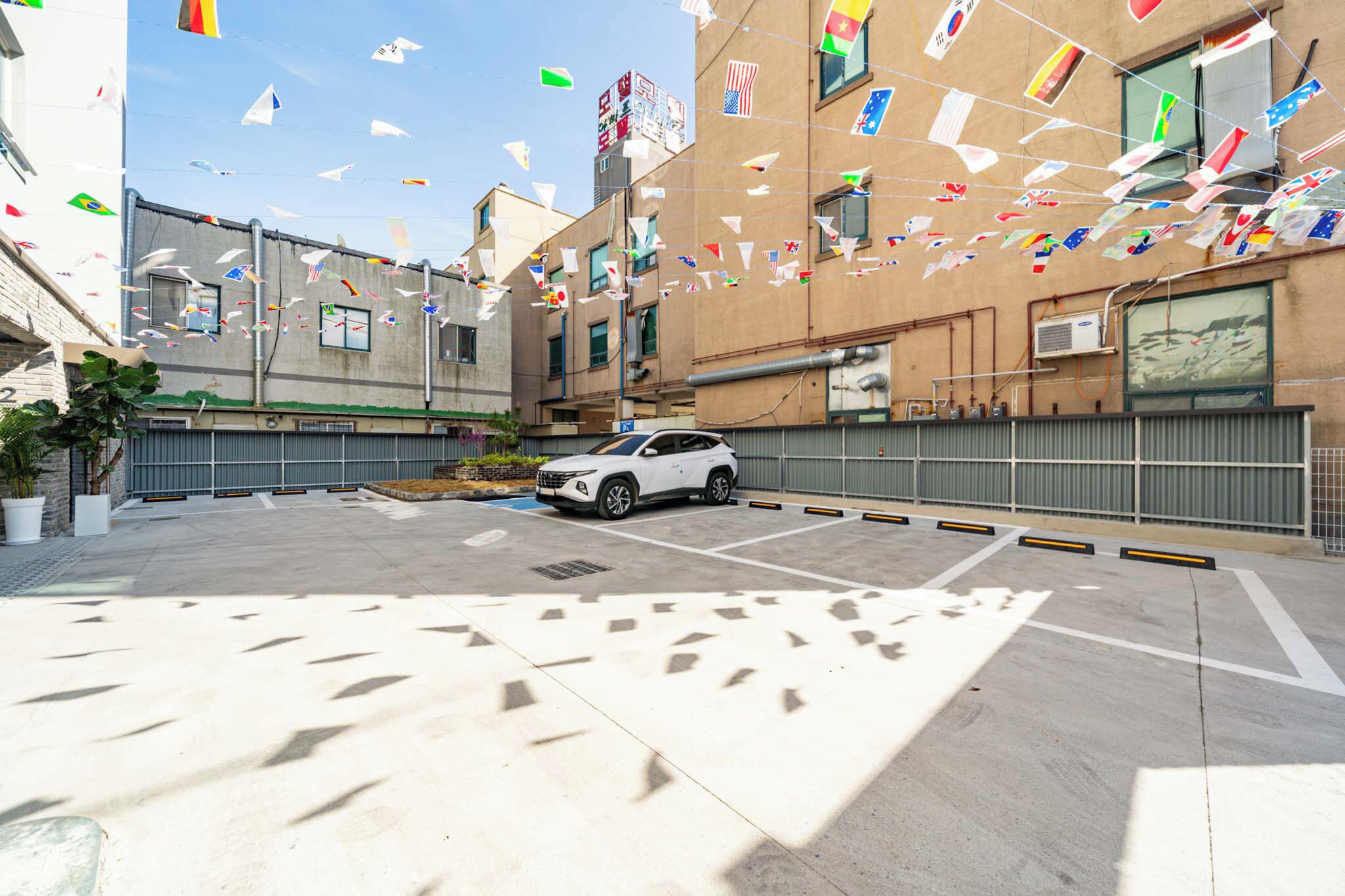 A parking area with a white SUV, surrounded by buildings with multiple flags.