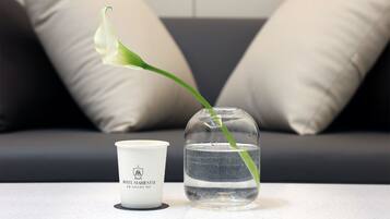 Una taza de cerámica blanca con un logotipo y texto, un florero de vidrio transparente con agua y una flor verde con tallo, y un sofá gris oscuro con cojines color beige.