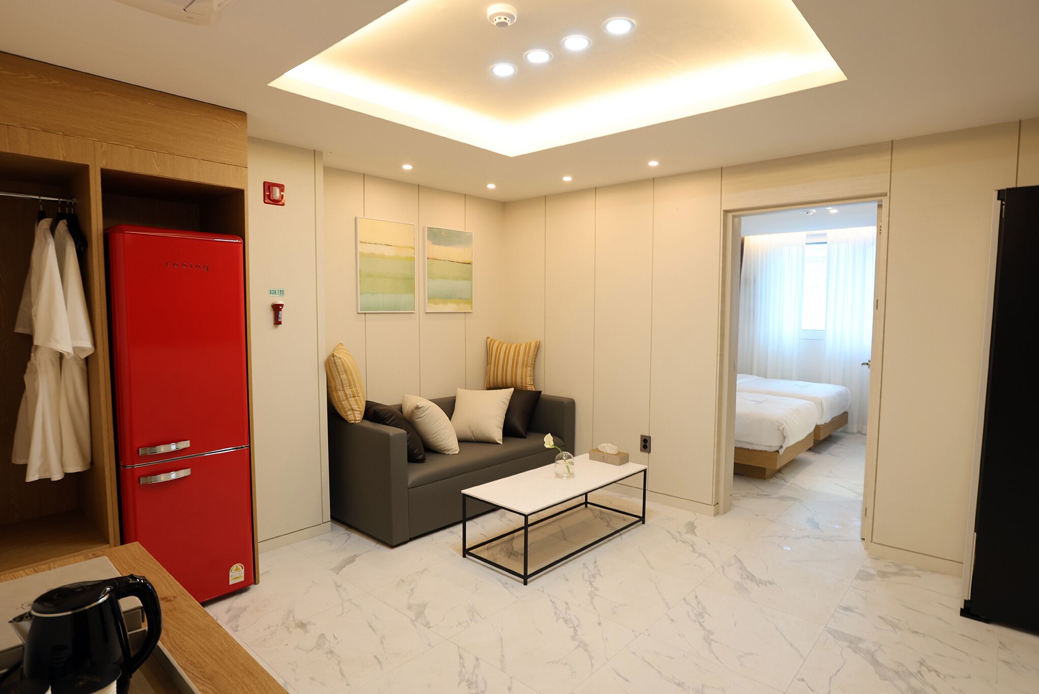 Habitación de hotel compacta con diseño moderno, que incluye un refrigerador rojo, un sofá con cojines, una mesa de centro y una cama con edredón blanco.