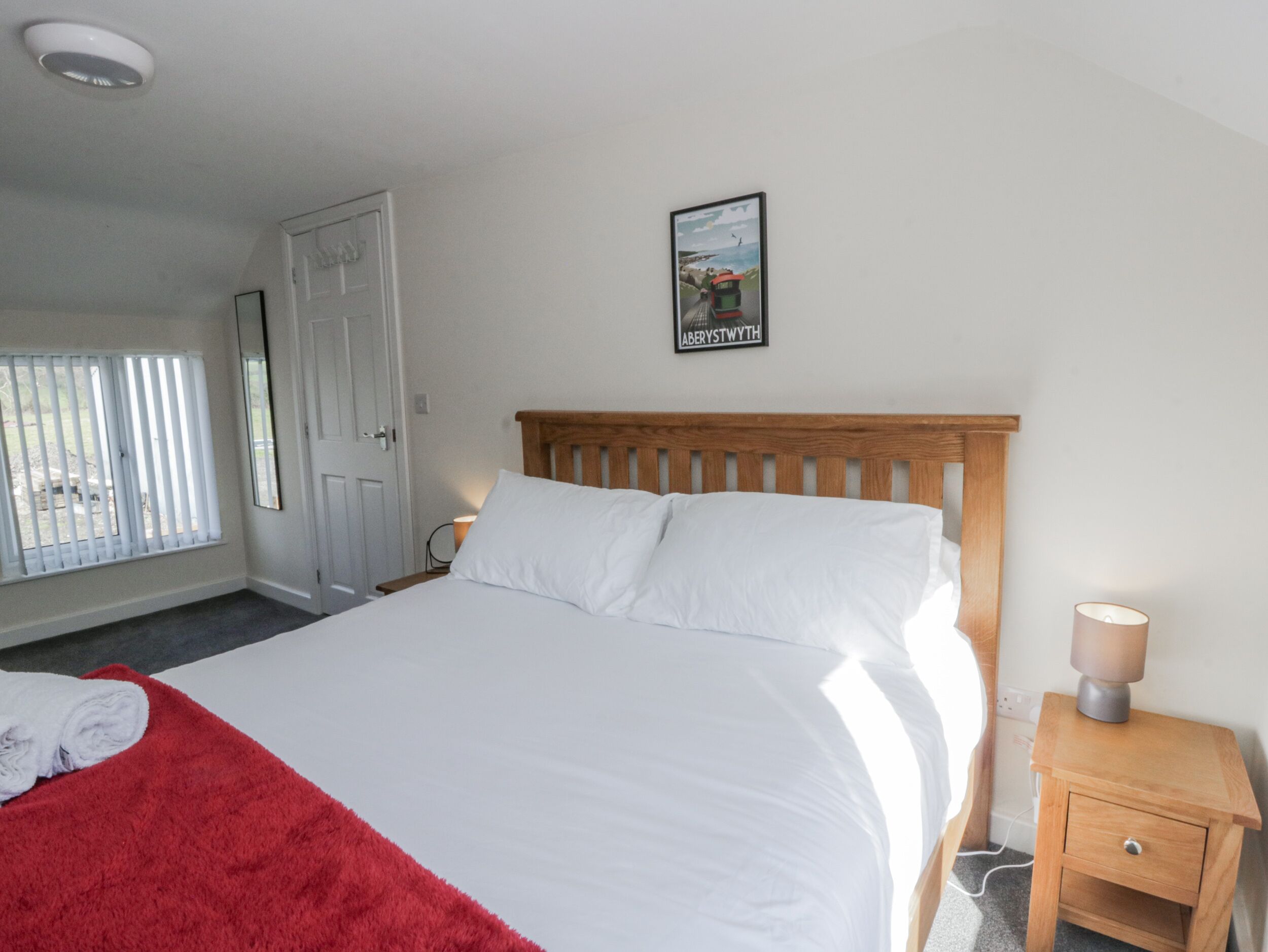 A neatly made bed with white linens, a wooden headboard, and a red throw blanket. There is a small bedside table with a lamp and a drawer. A framed picture hangs on the wall above the bed.