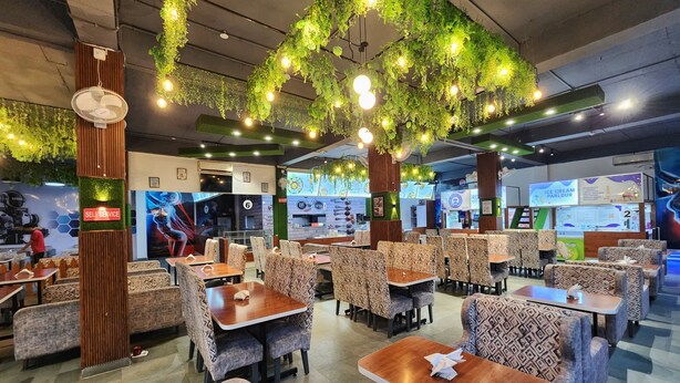 A restaurant interior with patterned seating, green hanging plants, and a self-service area. - Hotel Robo Nest (Balichauki)