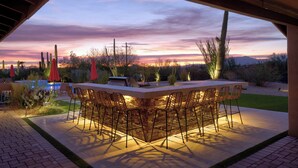 Un espace bar extérieur avec une piscine, entouré de cactus et de plantes, sous un ciel au coucher du soleil.